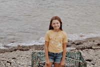 a young girl standing next to a basket of lobster traps on the beach